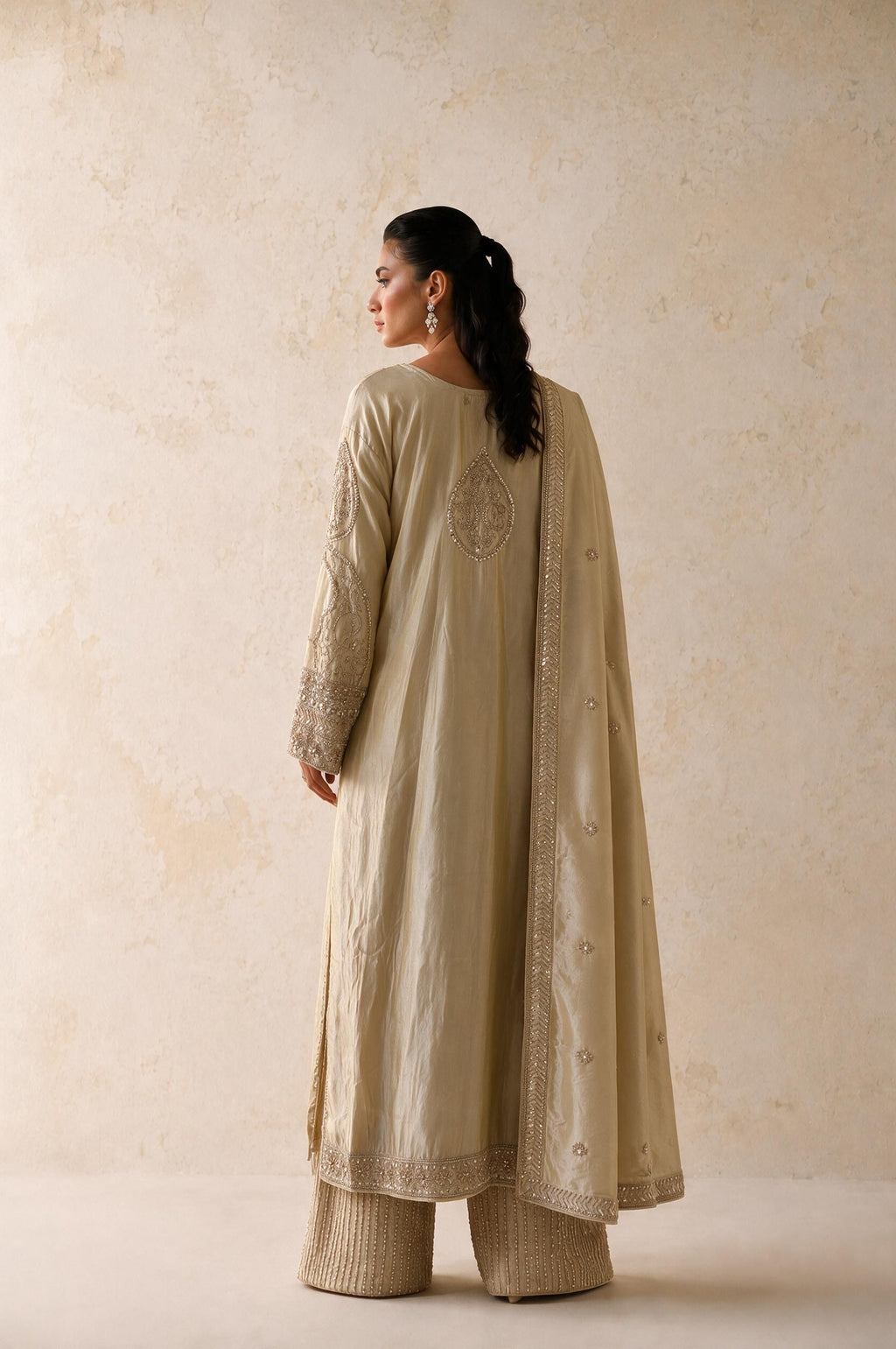 A woman wearing a beige traditional outfit with a long embroidered kurta and matching wide-leg pants, photographed from the back against a textured beige wall.