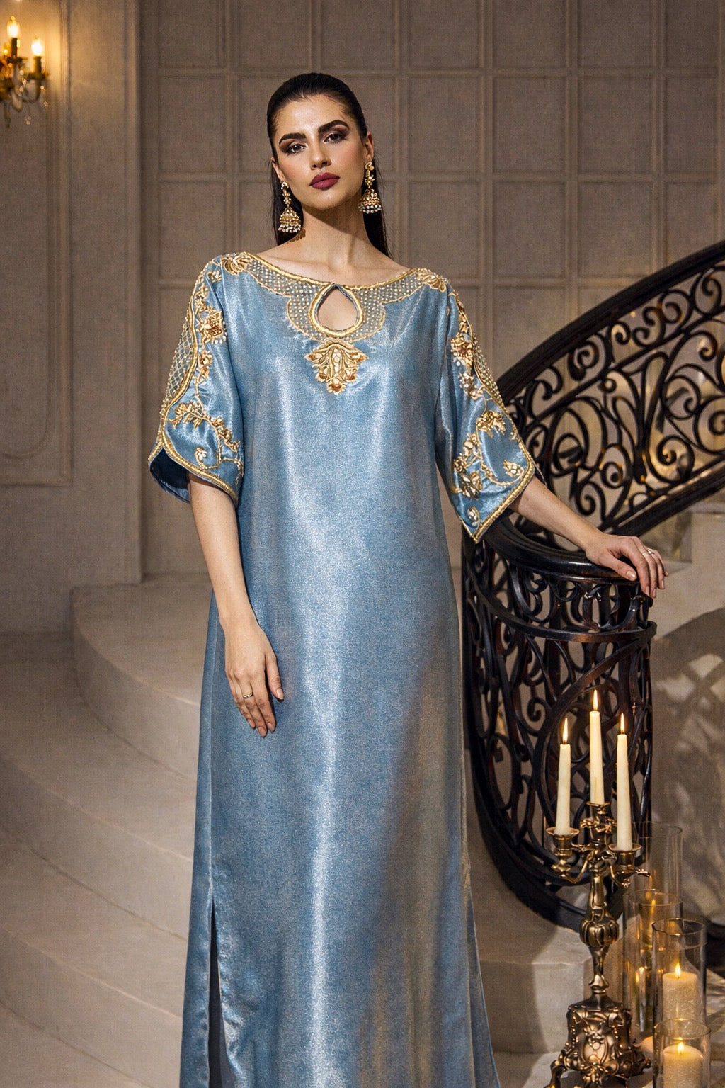 Woman in a blue dress with gold embroidery standing in an elegant interior setting.