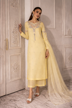 Woman in a light yellow traditional outfit with intricate designs, standing against a decorative wall.