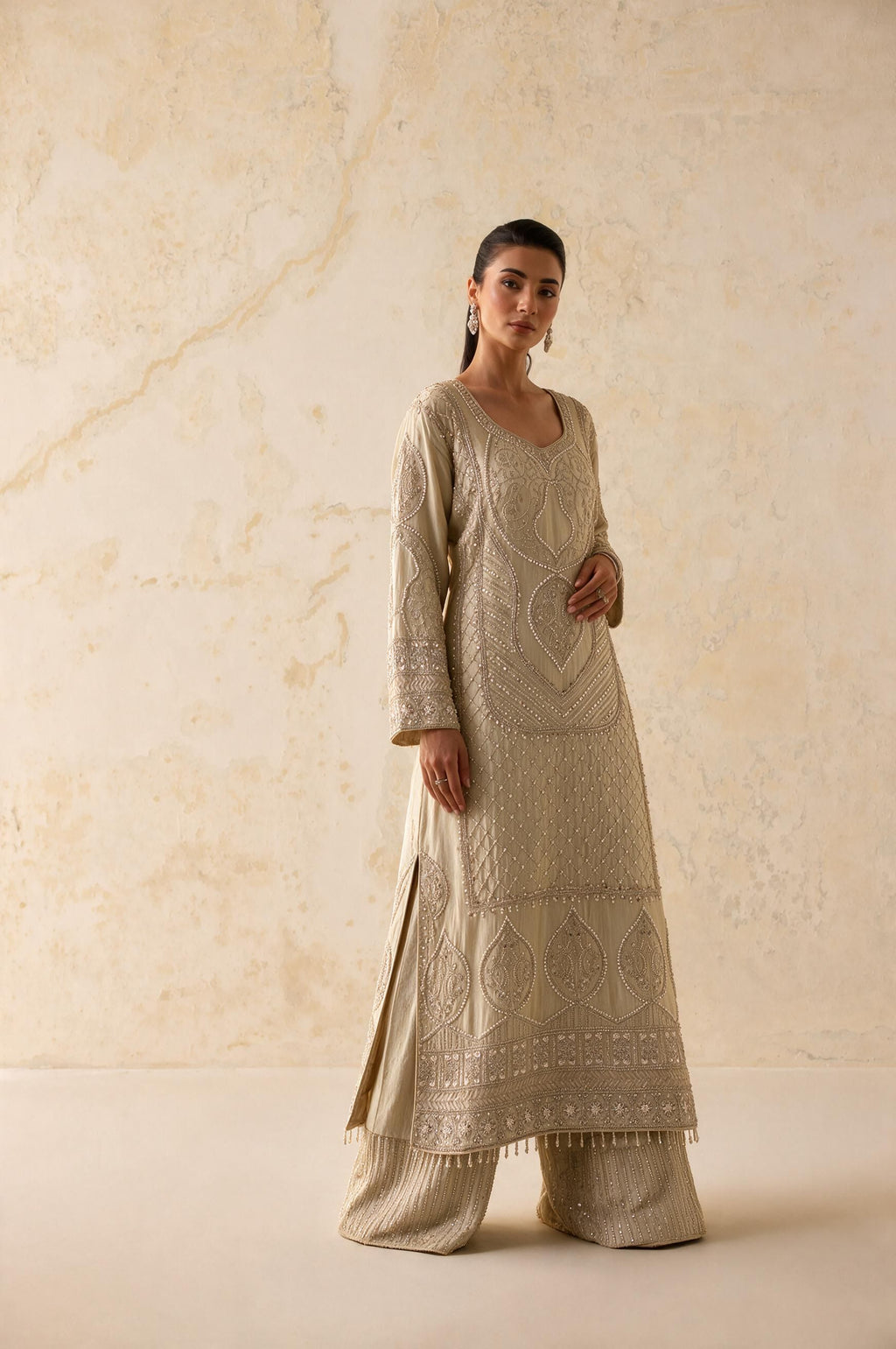 A woman modeling a beige embroidered kurta set with matching wide-leg pants against a textured beige wall.