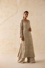 A woman modeling a beige embroidered kurta set with matching wide-leg pants against a textured beige wall.