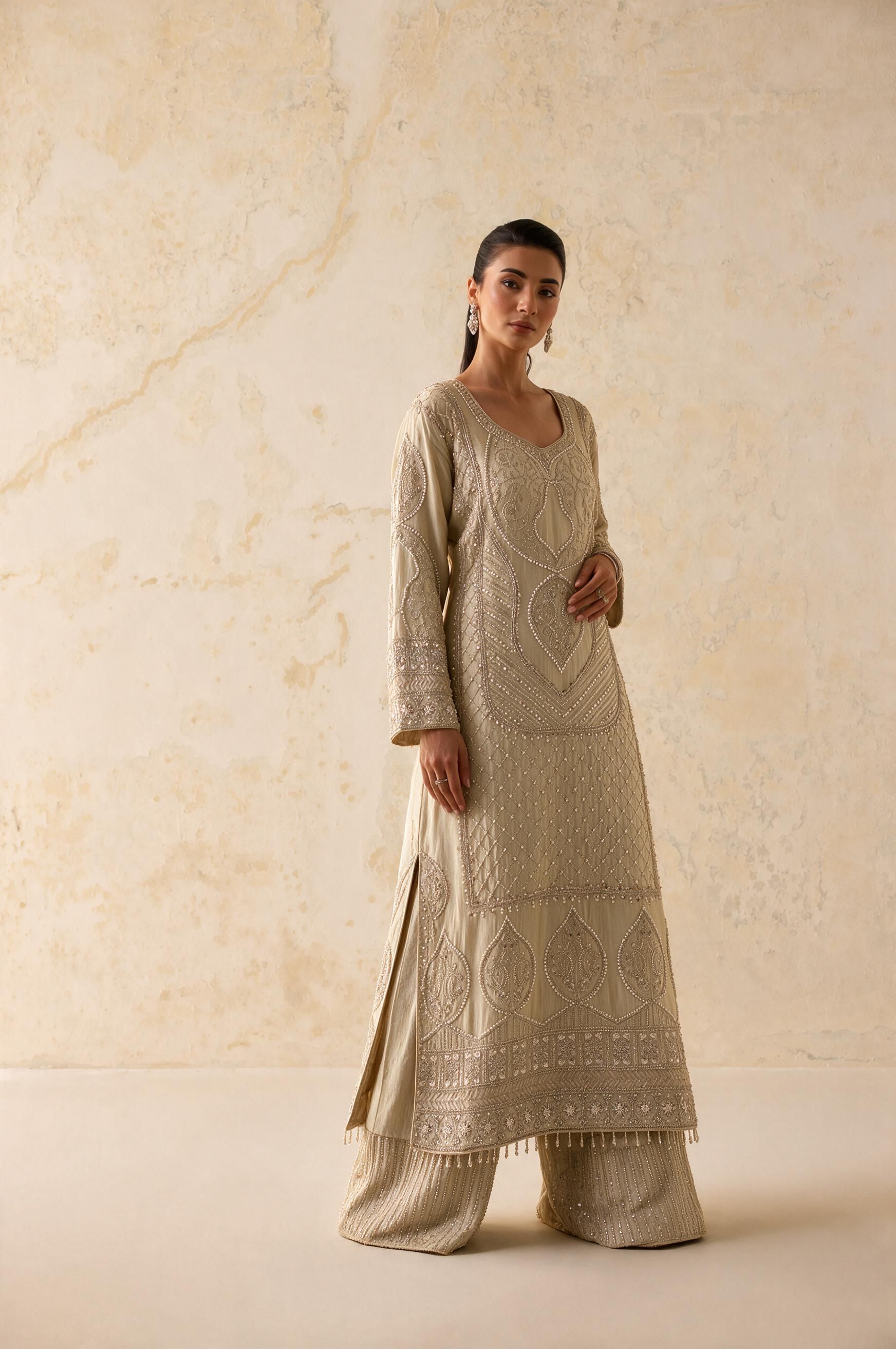 A woman modeling a beige embroidered kurta set with matching wide-leg pants against a textured beige wall.