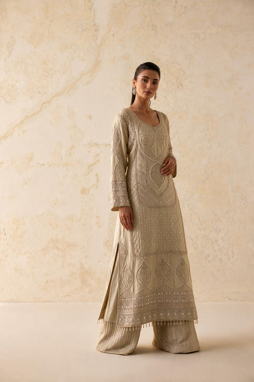 A woman modeling a beige embroidered kurta set with matching wide-leg pants against a textured beige wall.