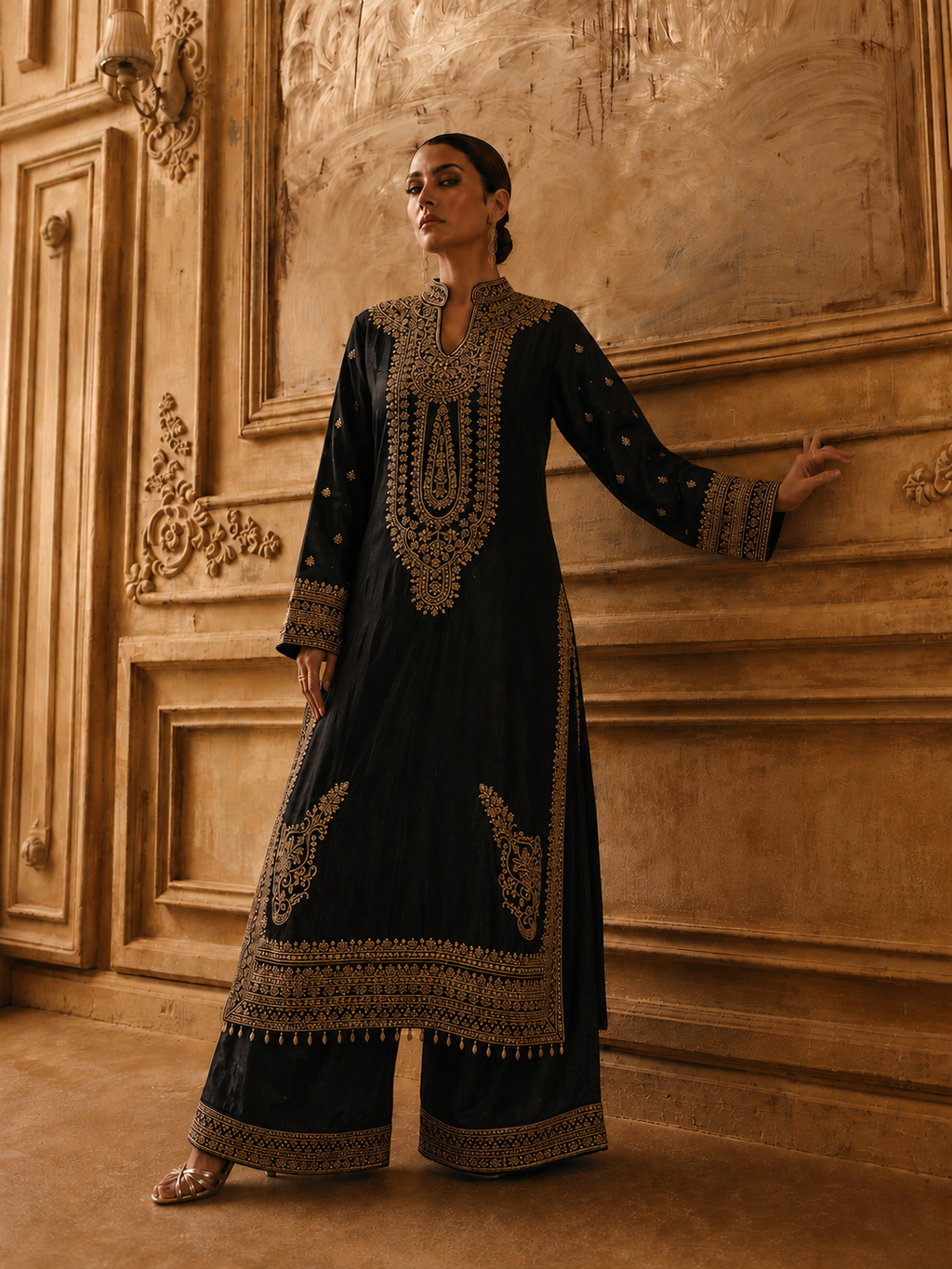 A woman models a black traditional long kurta with extensive gold embroidery, paired with matching wide-leg trousers, set against an ornate, warm-toned classical wall.