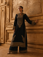 A woman models a black traditional long kurta with extensive gold embroidery, paired with matching wide-leg trousers, set against an ornate, warm-toned classical wall.