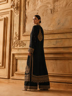 A woman models a black traditional long kurta with wide matching trousers, featuring ornate gold embroidery on the back, sleeves, and hem, set against a warm, classical stone wall.