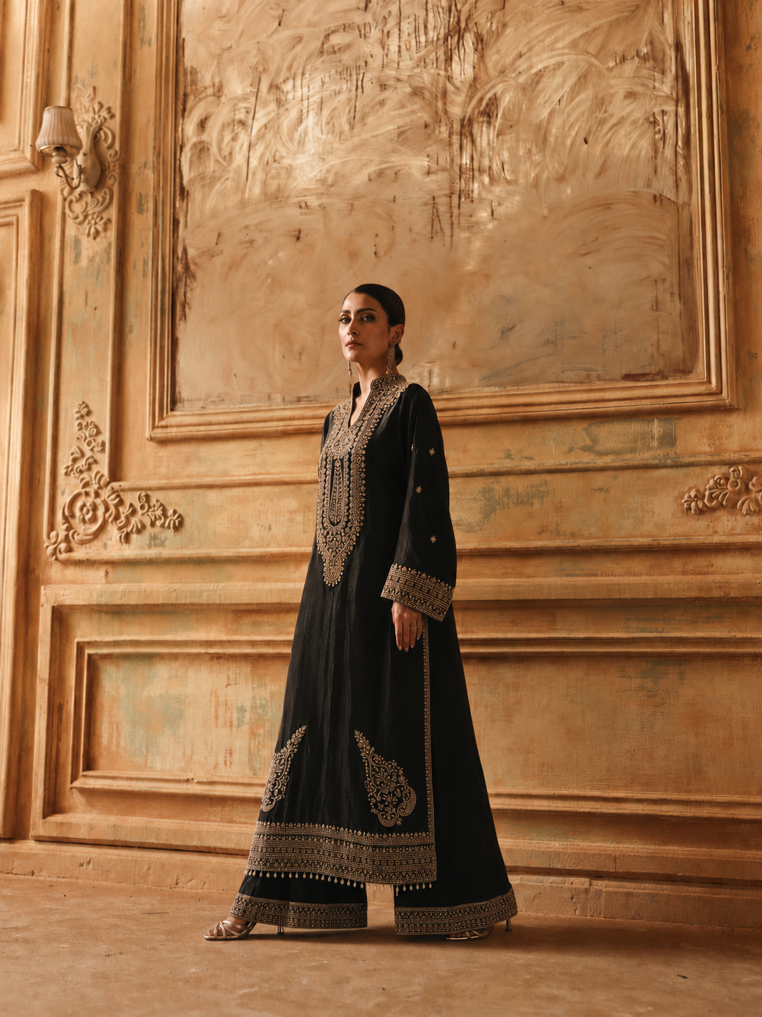 A woman stands in a grand interior wearing a long black traditional outfit with ornate gold embroidery and wide-leg trousers. The background features a large decorative wall panel in warm beige and brown tones.