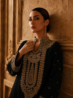 A woman wearing a black traditional outfit with elaborate gold embroidery, leaning against a textured warm brown wall.