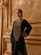 A woman modeling a black long kurta with elaborate gold embroidery on the front, collar, and cuffs, posed against an ornate beige wall.
