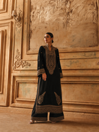 A woman stands in a grand interior wearing a long black traditional outfit with ornate gold embroidery and wide-leg pants. The background features warm beige plaster walls and decorative molding.