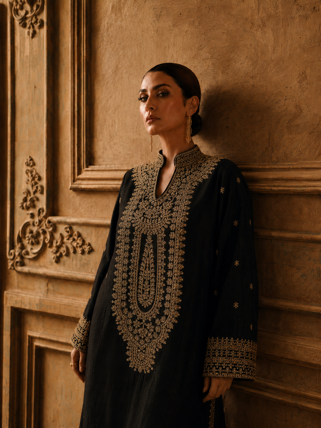 A woman wearing a black long kurta with ornate gold embroidery around the neckline, sleeve cuffs, and hem, leaning against a textured warm brown wall with carved paneling.