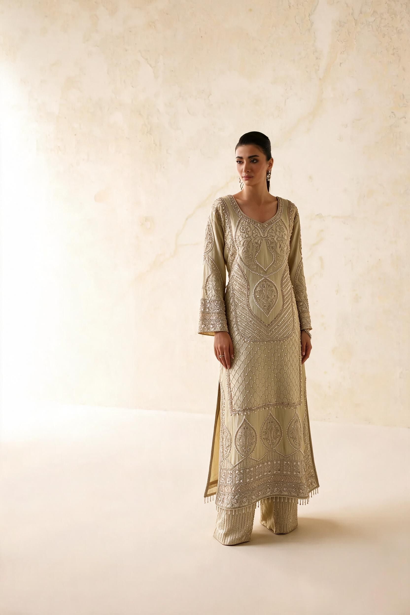 A woman modeling a traditional long beige embroidered kurta with matching wide-leg trousers, standing against a textured beige wall.