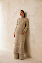 A woman wearing a beige embroidered kurta and matching wide-leg trousers stands against a textured beige plaster wall, looking off-camera.